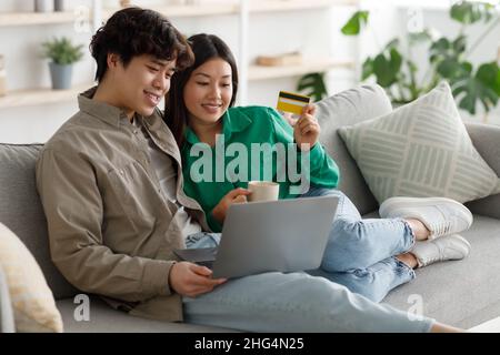 Happy Asian couple shopping online via laptop and credit card, ordering things in internet, sitting on sofa at home Stock Photo