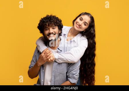 Lovely indian woman hugging her husband from behind, smiling and feeling happy on yellow studio background Stock Photo