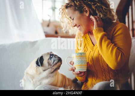 Smiling young adult woman have relax leisure at home drinking looking at her pug dog. MOdern female people with curly hair sitting on sofa Stock Photo