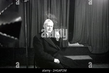 1950s, historical, a man, possibly an actor or performer in rehearsals, sitting in his overcoat by a stage and curtains on a break, having a cigarette, England, UK. Stock Photo