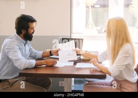 Side view of two multiethnic colleagues discuss financial data in report holding documents and charts sitting at desk in cafe by window. Stock Photo