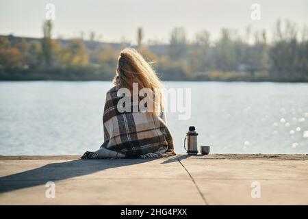 Woman relaxing alone by a river in the city wrapped in wool blanket enjoying nature and drinks tea from thermos. Picnic, rest, travel concept. Back vi Stock Photo