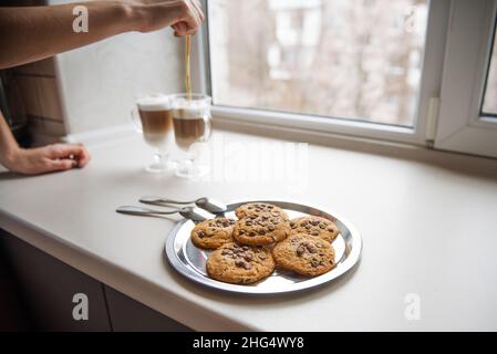 Young couple of millennials in black t-shirts hugging in the kitchen in the morning by the window. Man hugs woman who is making latte for breakfast at Stock Photo