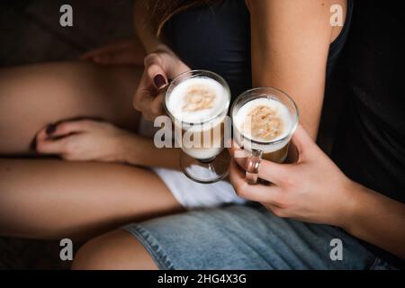 The hands of young couple hold glass cups with hot latte coffee. Lovers sit on the windowsill by the window, drink hot drinks in the kitchen at home i Stock Photo