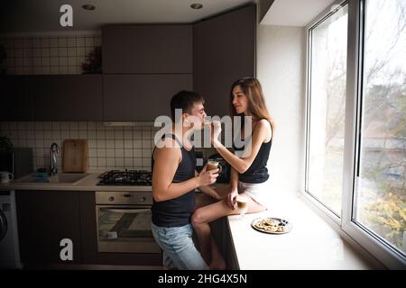 A young millennial couple is sitting on the windowsill near the window, drinking latte coffee with cookies in the kitchen at home in the apartment. Lo Stock Photo