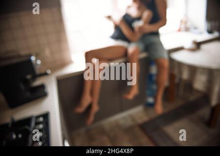Defocesed young millennial couple is sitting on the windowsill near the window, drinking coffee in the kitchen at home in the apartment. Lovers hug, k Stock Photo