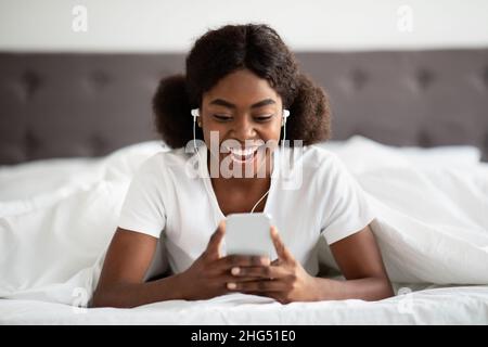 Happy young black woman laying in bed with smartphone, earphones Stock Photo