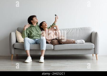 Millennial Asian couple turning on air conditioner, using remote control, suffering from summer heat at home, mockup Stock Photo
