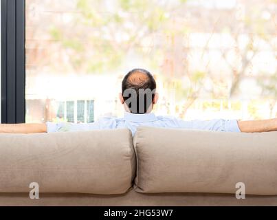 Rear view of a bald and carefree man sitting on a sofa at home and looking outdoors through the window at home Stock Photo