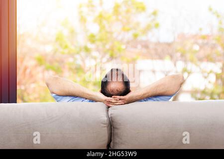 Rear view of a bald and carefree man on a couch at home and looking the green background outdoors through the window in the living room Stock Photo