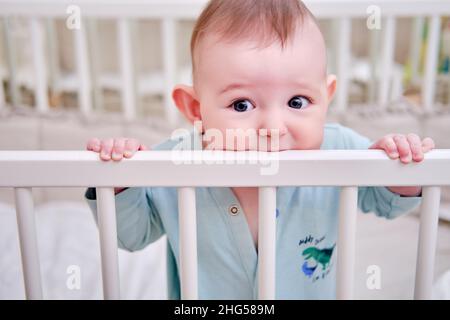 Baby gnaws at the edge crib during teething itching. Funny child scratching his teeth on the rail bed, age six months Stock Photo