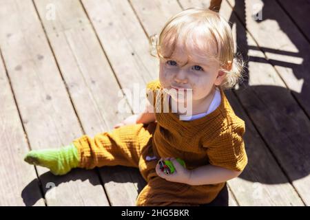 High angle view of a mischievous three year old toddler sitting on wooden decking holding lighters with a dirty face after playing in backyard. Stock Photo