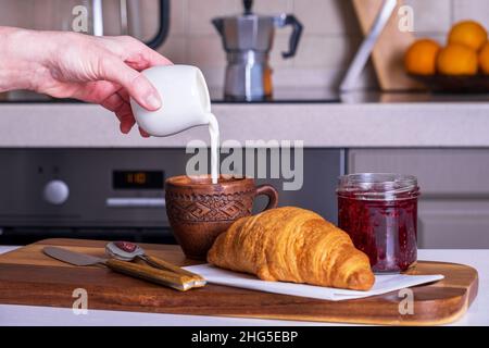 Fresh croissant, jam and cup of coffee pouring cream in woman's hand. Stock Photo