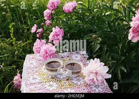 Tea in country style in summer garden in the village. Two cups of black tea on handmade crocheted vintage lacy tablecloth and blooming peony flowers i Stock Photo