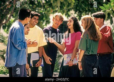 Teenager senior school students 15-17 years outdoors campus multicultural multi ethnic group of seven happy attractive teenage senior school college high school multicultural students in colourful t shirts relaxing talking and interacting outside in a sunny school college campus environment Stock Photo
