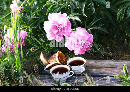 Tea in country style in summer garden in the village. Two cups of hot black tea and pancakes on wooden weathered boards and blooming pink peony flower Stock Photo