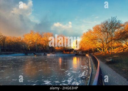 Winter city park at sunset and a frozen lake with trees without leaves Stock Photo