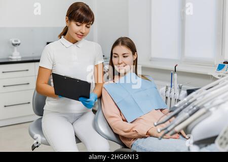 Friendly dentist with tablet explaining treatment to patient during teeth examination. Modern dental clinic. Concept of professional oral care, medical consultation in stomatology office. Stock Photo