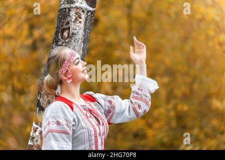 Portrait of young beautiful smiling woman in folk traditional slavic clothes greets the sun at nature in autumn forest. Stock Photo