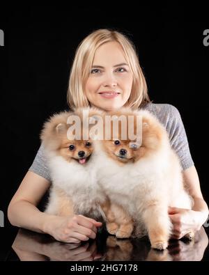 Smiling pretty woman holding two cute puppies of pomeranian spitz breed dog. Owner with her friends pets at black background. Stock Photo