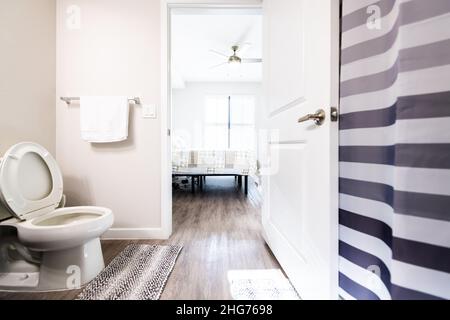 Empty master bath bathroom interior in new modern luxury apartment home house white toilet seat up rugs open door to bright bedroom and shower curtain Stock Photo