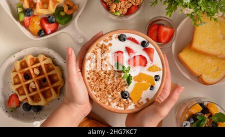 Top view, A female hands holding a smoothie yogurt bowl with fresh berries and granola over table. Stock Photo