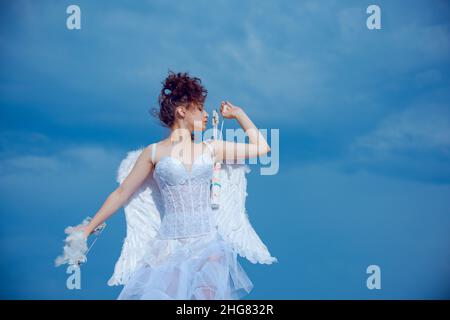 Cute girl cupid with wings aiming with bow and arrow on a sky background. Valentines day concept. Angel teen in heaven. Stock Photo