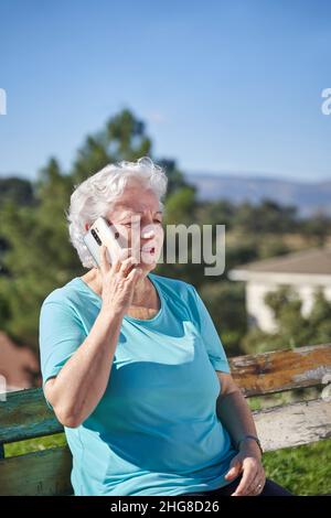 Old female with wrinkled face and gray hair sitting on bench and talking on mobile phone in sunny day under blue sky Stock Photo