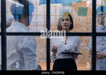 20s Asian company female manager brainstorming in the meeting room while having sticky notes on glass in office. She's holding a tablet while working. Cooperation and teamwork concept Stock Photo