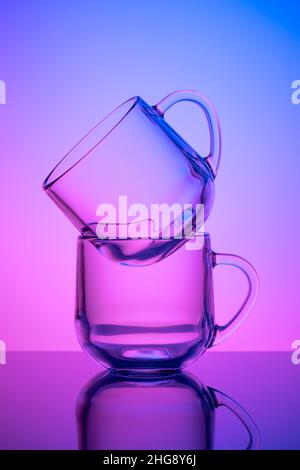 Two empty tea cups on a pink and blue neon background. Drinking glass. Dark silhouette, night illuminated effect. Glassware. Transparent tableware, di Stock Photo