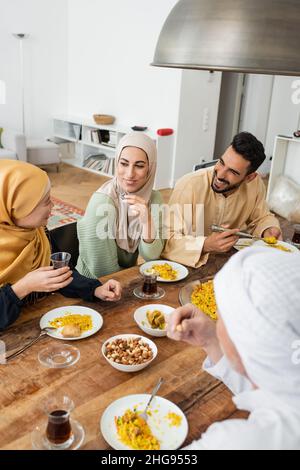 mature asian woman holding glass of tea while talking to muslim family during dinner