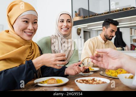 cheerful asian woman holding cup of tea during dinner with muslim family