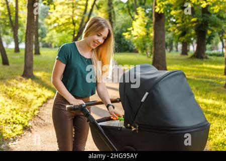 Young blonde woman walking with black stroller in summer park. Happy mother with baby in pram outdoors.  Stock Photo