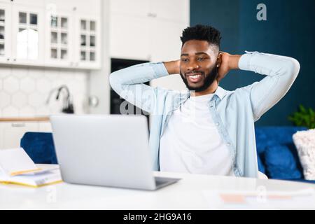 Smiling man sitting at the laptop, holding hands behind head, relaxing, looking at the screen, while working in comfortable office. Man did the job productively and got praise Stock Photo