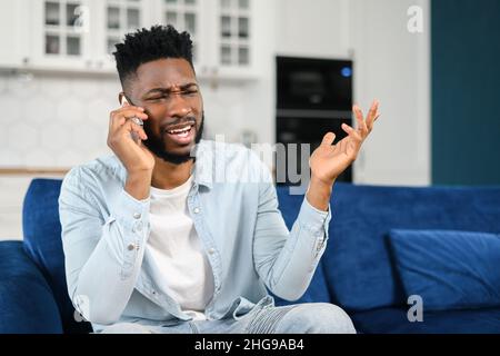 Angry stressed multiracial man having unpleasant phone conversation, talking on the smartphone and arguing, sitting on the sofa and speaking with frustrated face expression Stock Photo