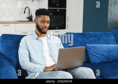Optimistic confident dark-haired handsome guy using laptop for work. Multiracial freelancer man sitting at the sofa and looking at the screen, smiling friendly Stock Photo