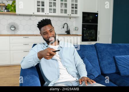 Happy young multiracial man holding tv remote control while sitting on a couch at home and relaxing at the weekend. Stock photo Stock Photo