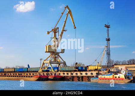 River port crane loading open-top gondola cars on sunny day. Empty river drag boats or barges moored by pier, Empty cars ready for loading. Stock Photo