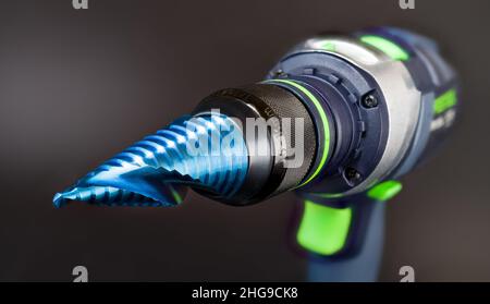 Blue nano coated cone step drill bit clamped in cordless screwdriver on gray background. Sharp tool to drilling varied hole size at metal or wood work. Stock Photo