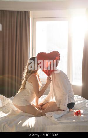 Beautiful young couple at home. Hug, kiss and enjoy spending time together while celebrating Valentine's Day on the bed with a red heart shaped Stock Photo