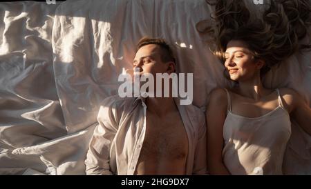 Romantic morning at the hotel. A man and a woman in white clothes and on white sheets of a warm hotel room lie in bed. Love, smile, honeymoon. Stock Photo