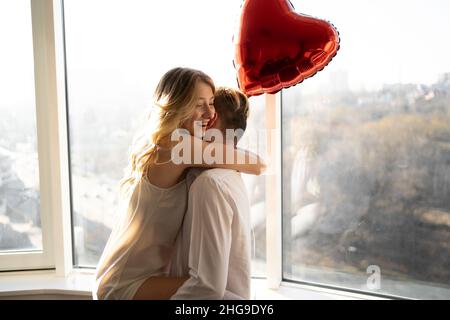Beautiful young couple at home. Hugging, kissing and enjoying spending time together celebrating Valentine's Day with a heart shaped balloon. Stock Photo