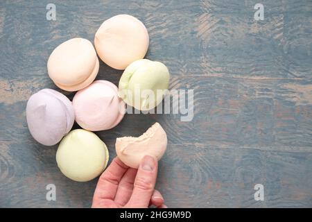 Round multi-colored marshmallows in a pile and a hand holding a bitten half of a marshmallow on a blue wooden background, top view copy space. Delicio Stock Photo