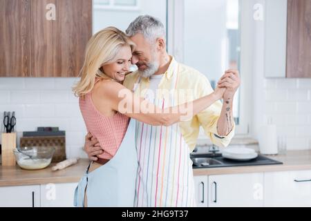 Tattooed mature man in apron dancing with smiling wife in kitchen Stock Photo