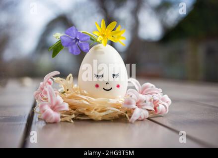 egg with a cute face painted with a smile and closed eyes is decorated with fresh spring flowers. creative ideas for decor. happy easter holiday. Fest Stock Photo