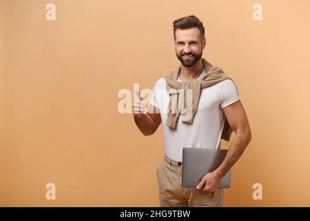 Overjoyed excited man with beard showing big finger and holding laptop while looking at camera with happy smile. Indoor studio shot isolated on orange background Stock Photo