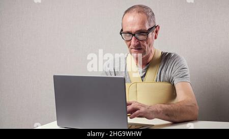 Senior man with fixating bandage works on laptop at desk Stock Photo