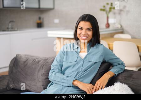 Carefree multiracial woman resting on the comfortable couch at home, while feeling happy. Charming woman looking at camera and smiling while resting at home Stock Photo