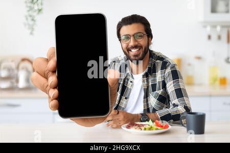 Handsome Indian Man Showing Blank Smartphone At Camera While Sitting In Kitchen Stock Photo
