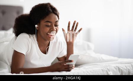 Cheerful black woman laying in bed, having video chat Stock Photo